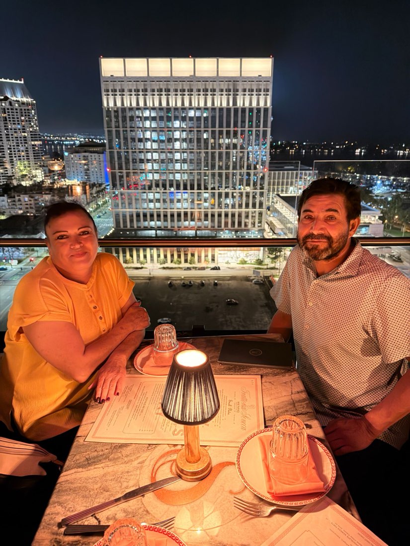 Mundo and his wife smiling at dinner, with the San Diego skyline behind them